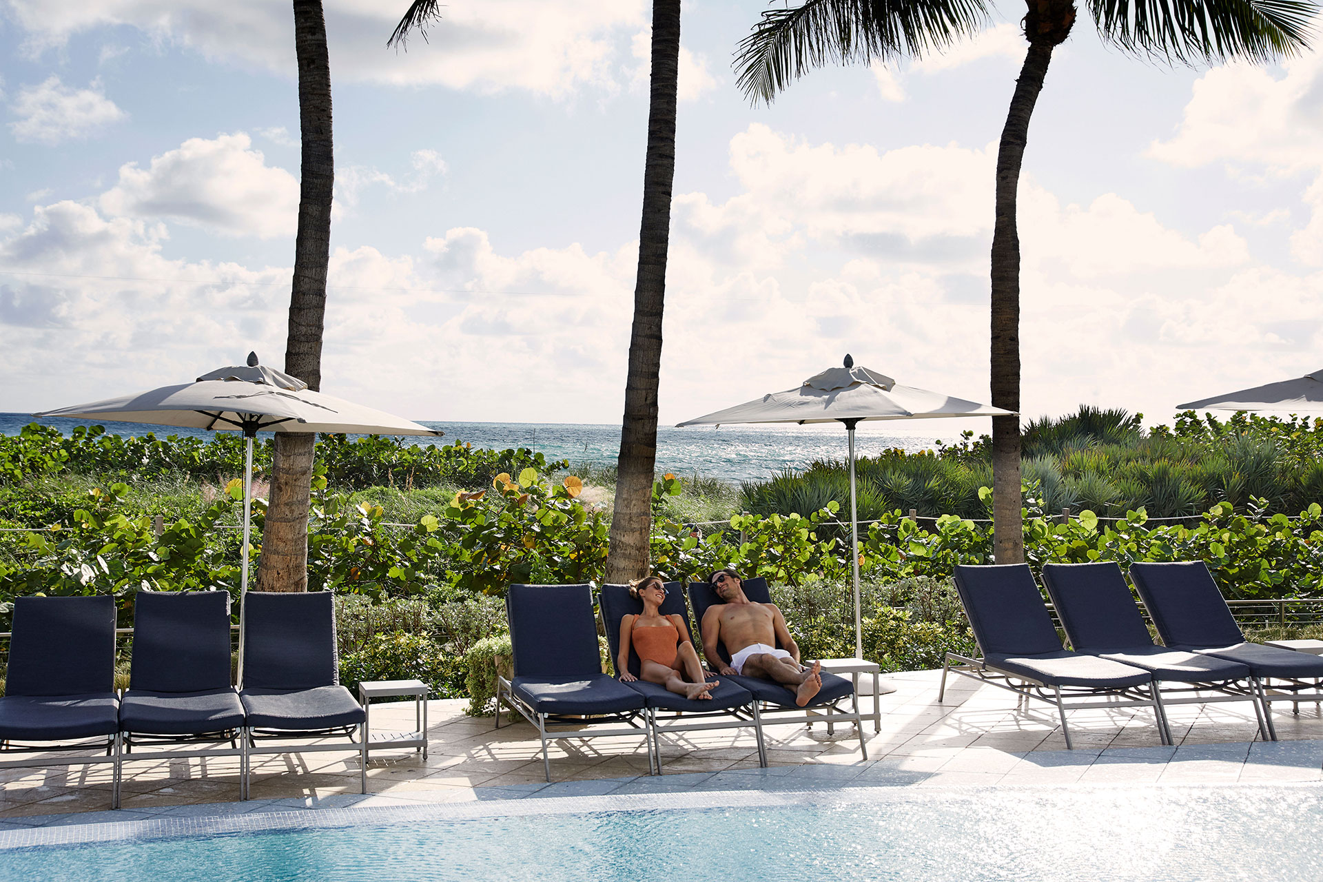 a man and a woman in swimwear lounge of blue pool chairs next to a pool. They are under a white umbrella. In the background is greenery and palm trees. The ocean peaks out in the distance.