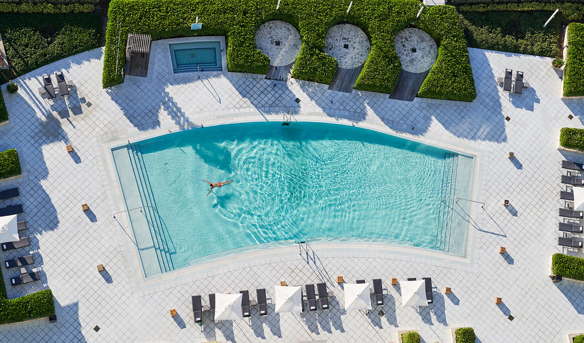 aerial image of a pool surrounded by white tile and lush green manicured bushes. Off to the side is a hot tub. There are gray lounge chairs adn white umbrellas lining the edge of the pool.