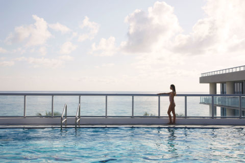 a woman in a bathing suit stands looking out over a balcony. There view is of the ocean and the resort grounds. In the foreground is a long swimming pool