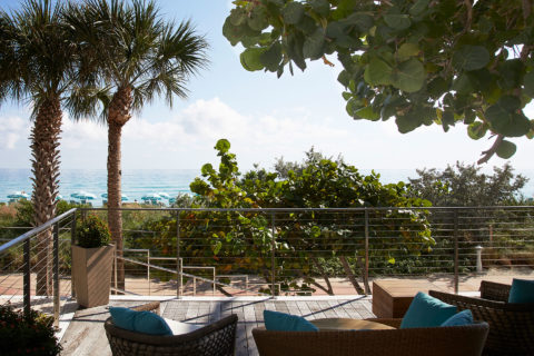 Image of an outdoor seating area. There are woven chairs and couches with blue throw pillows on a balcony. The balcony is surrounded by greenery such as palm tress and bushes. There is a view of the ocean and the beach chairs and umbrellas that line the sand.