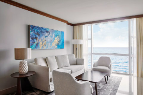 a sitting room in a suite with an ocean view. There is a light colored couch against the wall with a blue painting overhead. There are two chairs with the same fabric on either side of the couch circling a dark wood table.