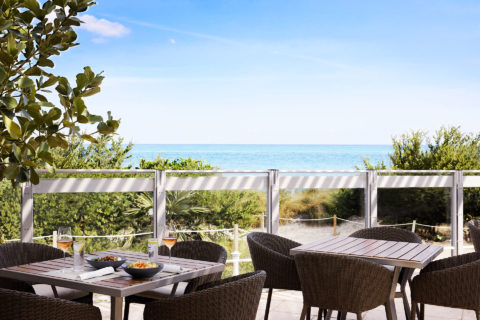 photo of an outdoor dining area. The chairs are woven brown and the tables are a dark wood. At on table there is a glass of rose at each place setting and there are two gray bowls of food in the middle of the table. The outdoor seating area overlooks the sand dunes and the ocean