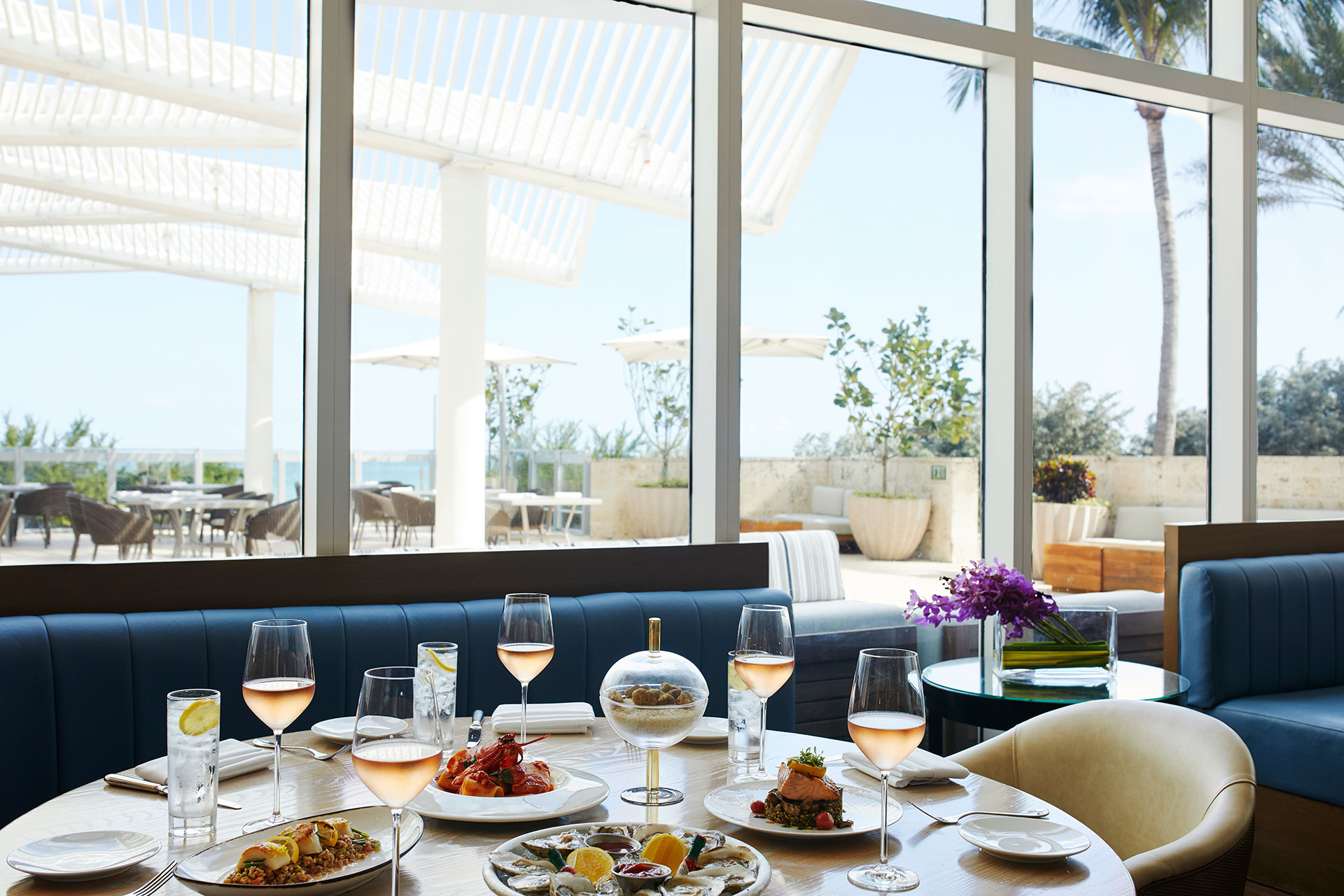 an interior shot of a dining room. There are full length windows that give a view of an outdoor dining area. The seating is plush, blue benches and white leather chairs. The circle table is full with rose glasses at each place setting and multiple dishes in the middle of the table.