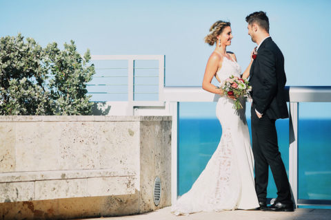 a wedding portrait. A man and a woman face each other, smiling. The woman is resting her had gently on the man's chest. The woman wears a slim, spaghetti strap lace dress and the an wears a black suit. The woman is holding a bouquet of red, pink and white flowers and the man wears a red rose on his chest. They are resting against a glass balcony and there is greenery next to them and a view of the ocean in the background.