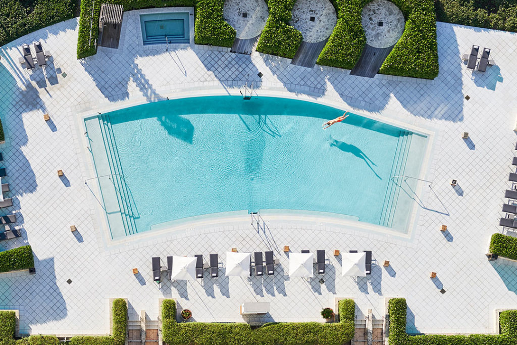 Aerial view of a pool with a person swimming, surrounded by lounge chairs and hedges.
