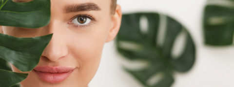 close up shot of a woman with minimal makeup and her hair pulled back. There is a palm leaf covering half of her face. There are more palm leaves in the background of the photo
