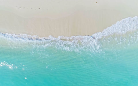 aerial shot of a wave crashing on the sand. The water is crystal clear light blue and the sand is a light beige. there are faint traces of footprints in the sand
