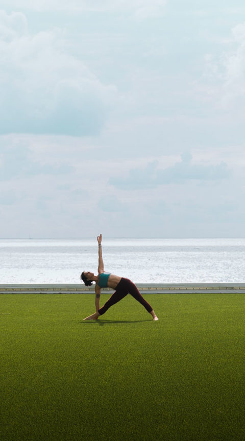 a woman is performing a yoga pose on a bright green lawn overlooking the ocean. She is wearing dark leggings and a bue sports bra. Her hand is reaching for the sky and she is looking up.