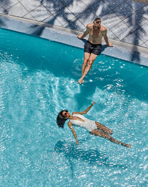 lady in a white suit floating in the blue pool water, man sitting on the pools edge