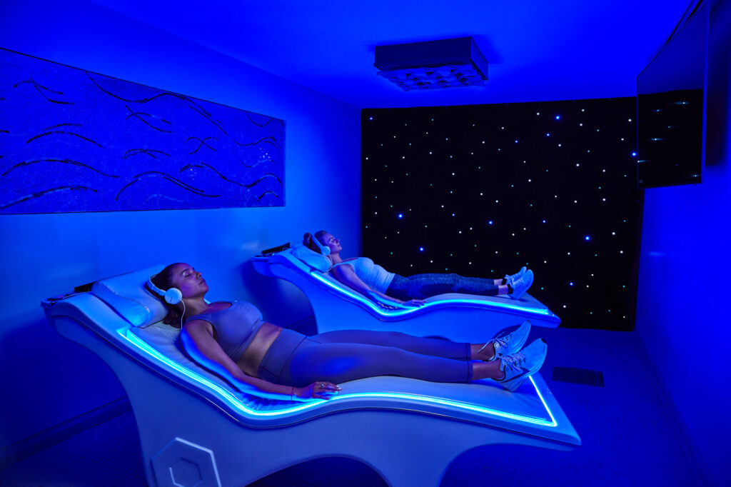 Two women relax in reclining chairs with blue lights and headphones in a spa-like room with a starry night ceiling.