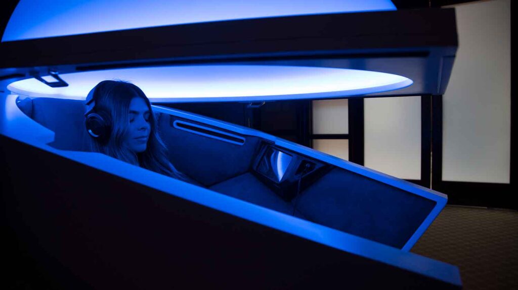 A woman wearing headphones lies in a pod emitting blue light.