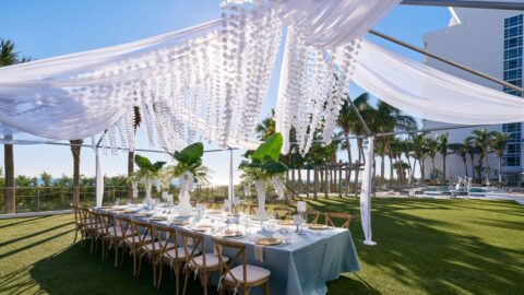 A long table set for a wedding celebration in the center of a tent with white curtains and flowers draping over